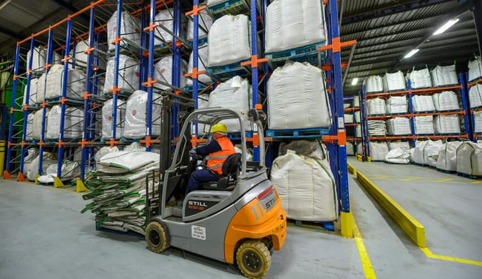 A worker in safety gear lifts a stack of items with an electric forklift in a large factory interior