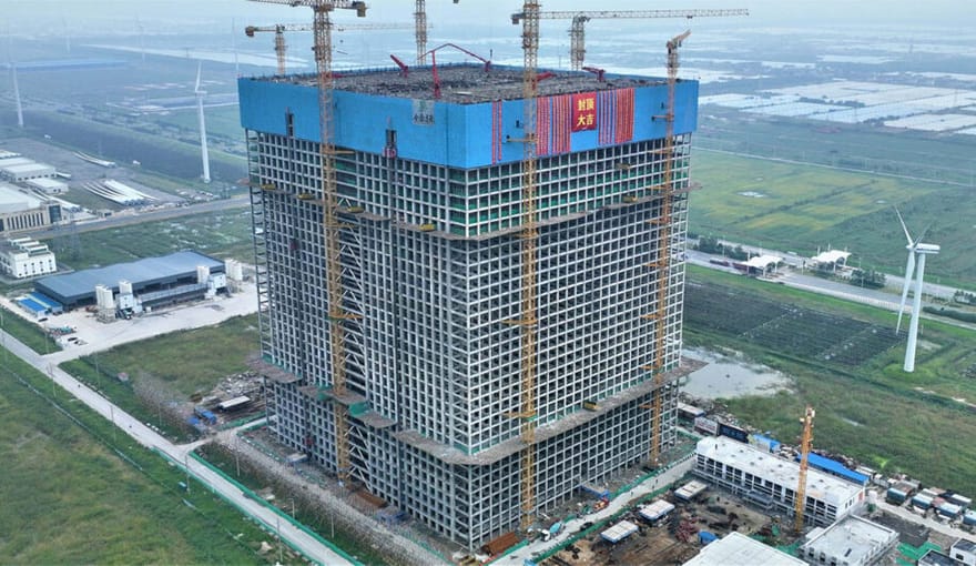 A large, tall, rectangular facility in a rural area. Wind turbines are seen in the background