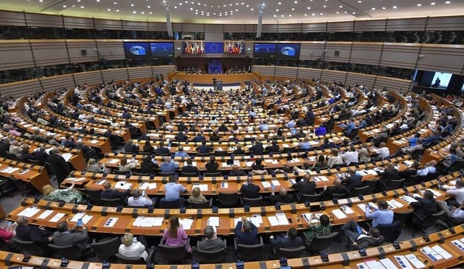A large legislative chamber full of people sitting at concentric rows of desks looking at a speaker on the main stage