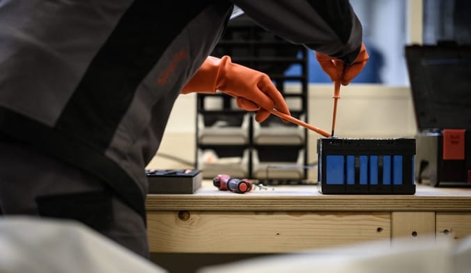 A person wearing orange gloves repairs a small battery on a workbench using two orange tools