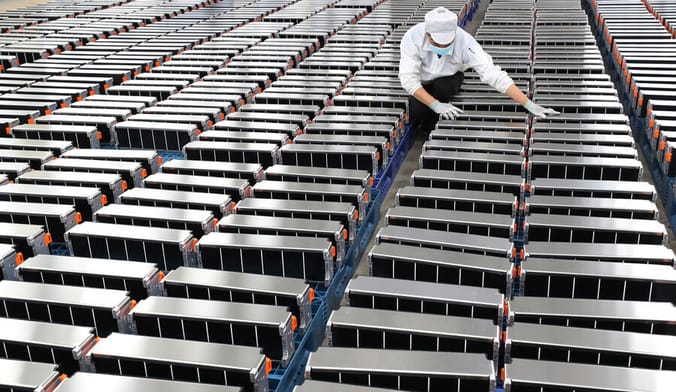 A factory worker in gloves, a white cap and mask stands amid rows of hundreds of EV batteries