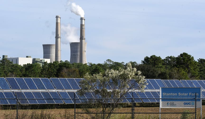 In the foreground is a large solar array. In the background are the stacks of a conventional fossil power plant
