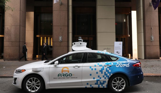 A small white and blue sedan with a camera affixed to the top, with the words Ford, Argo and self-driving test vehicle