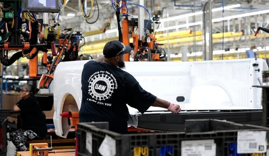 A Black man wearing a UAW t-shirt with his back to the camera works in a factory