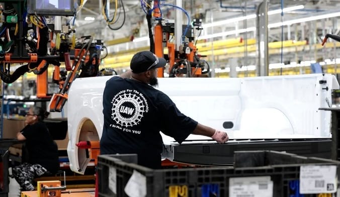 A Black man wearing a UAW t-shirt with his back to the camera works in a factory