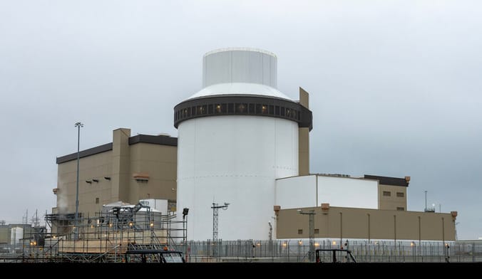 A large beige and white nuclear reactor facility surrounded by fencing under a cloudy gray sky