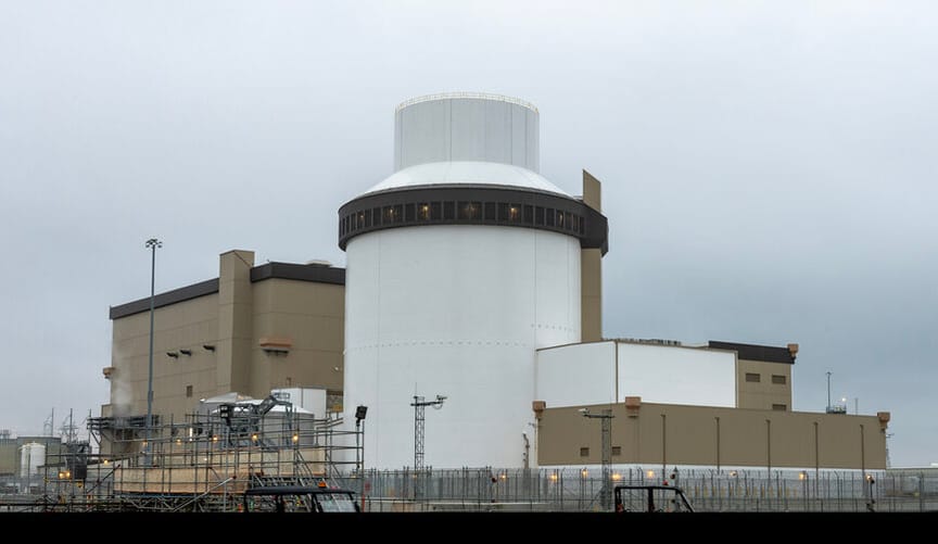 A large beige and white nuclear reactor facility surrounded by fencing under a cloudy gray sky