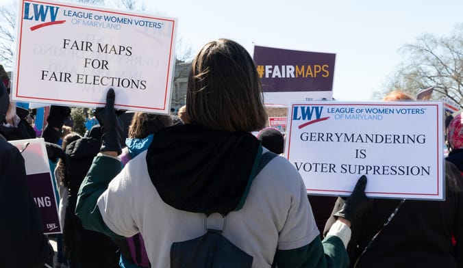 A protestor holds signs that say fair maps for fair elections and gerrymandering is voter suppression