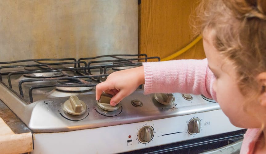 A young girl with light brown hair wearing a pink sweater turns a knob on a gas stove