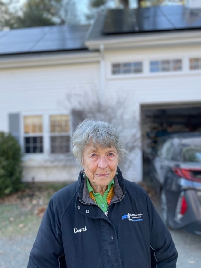 An elderly woman with gray short hair stands in front of a white house with solar panels on the roof