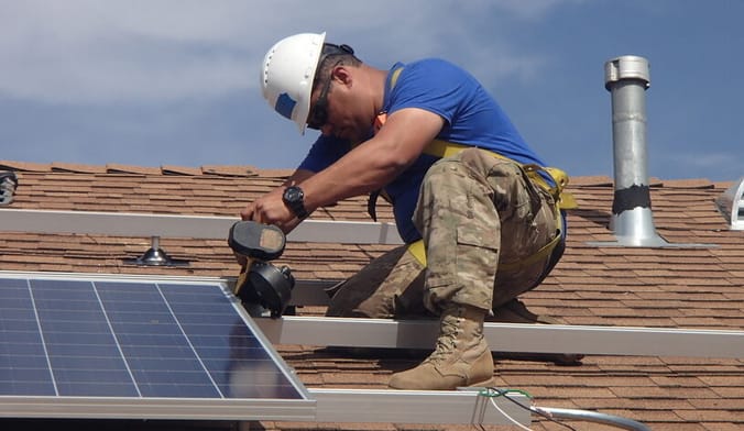 A man in a white hard hat, blue shirt and camo pants installing a solar panel on a roof