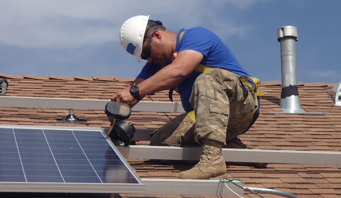 A man in a white hard hat, blue shirt and camo pants installing a solar panel on a roof