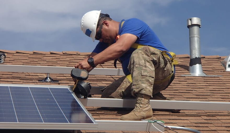 A man in a white hard hat, blue shirt and camo pants installing a solar panel on a roof