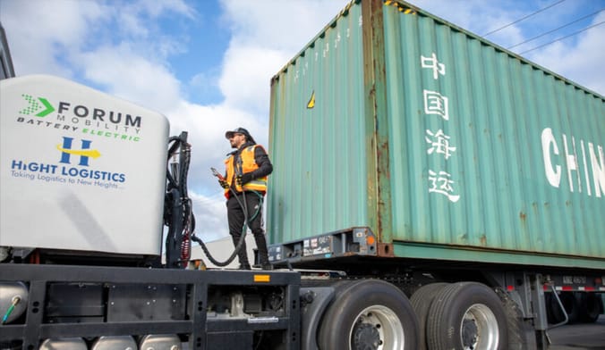 A man wearing a baseball cap and an orange safety vest stands on a platform next to a semi trailer