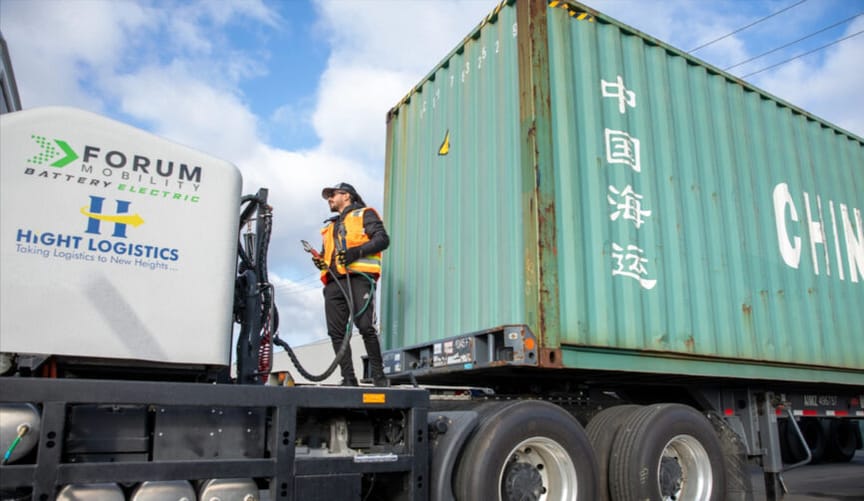A man wearing a baseball cap and an orange safety vest stands on a platform next to a semi trailer