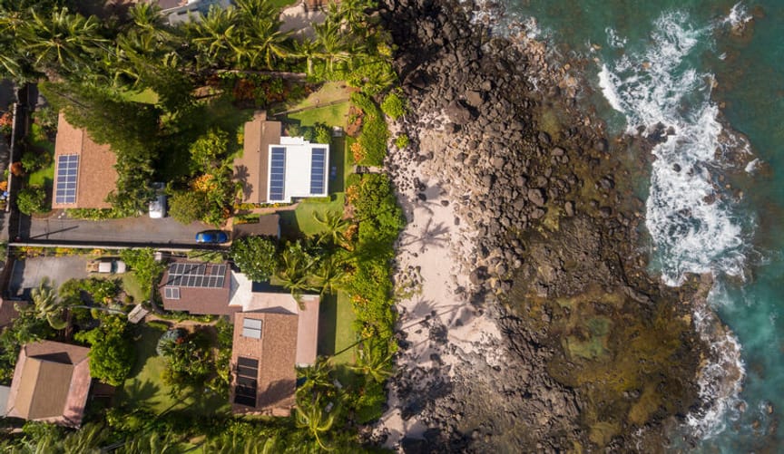 An overhead view of a small cluster of homes with solar panels on the roofs next to a turquoise ocean and dark sandy beach