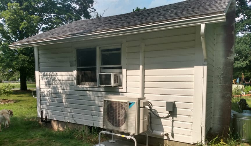 A small beige cottage on a wooded lot with a new heat pump installed on the exterior