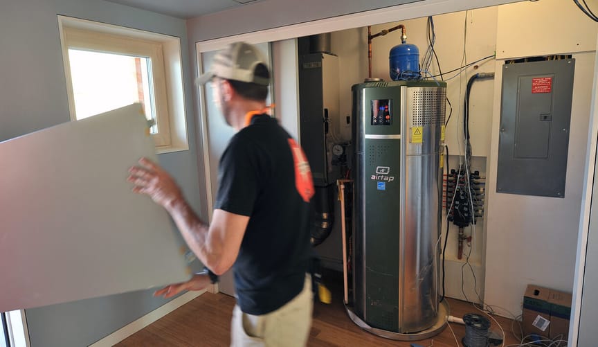 A man holding a white rectangular object walks past a closet with a large silver water heater installed