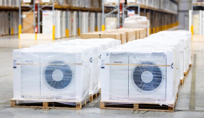 Two rows of white metal heat pumps wrapped in transport packaging sit on pallets on the floor of large warehouse