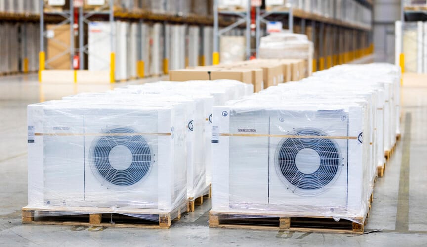 Two rows of white metal heat pumps wrapped in transport packaging sit on pallets on the floor of large warehouse