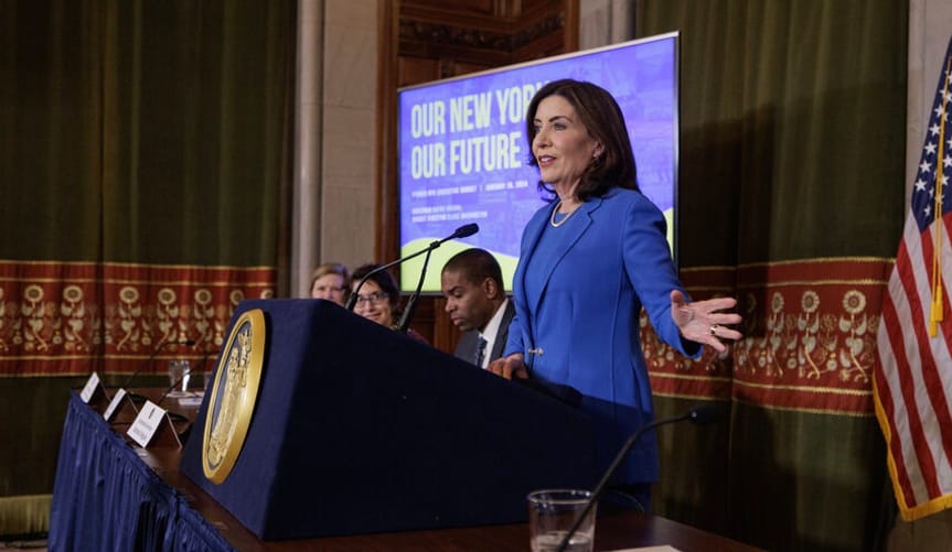 A woman with a light skin tone and shoulder length brown hair wearing a blue jacket speaks and gestures behind a lectern
