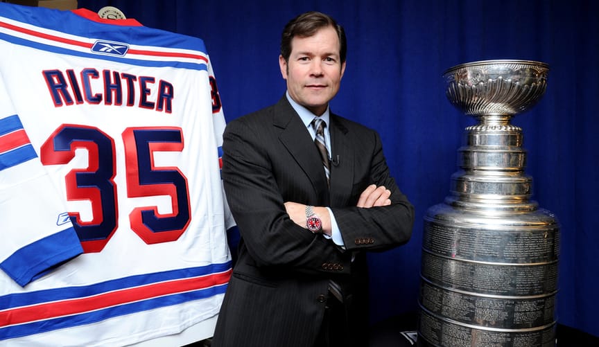 A man with light skin and dark hair wearing a dark business suit stands by a large trophy and a jersey that says Richter 35