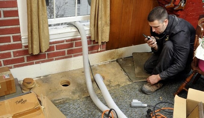 A man in protective work gear crouches on the floor of a home next to a long tube installing insulation into a crevice