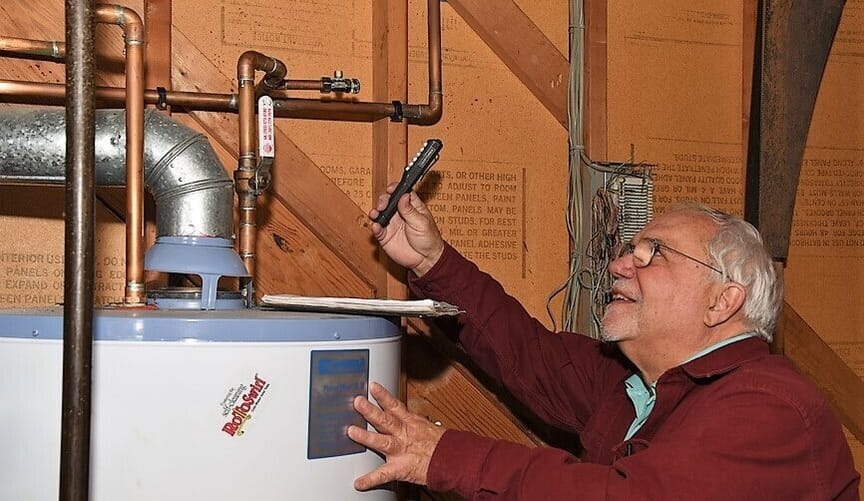 A smiling white man with glasses and white hair uses a tool to assess a hot water heater