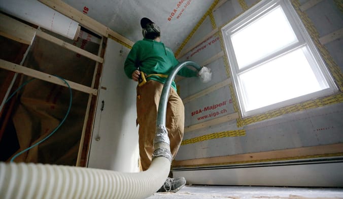 A man in work clothes and wearing a protective mask holds a large hose that's connected to an interior wall of a house