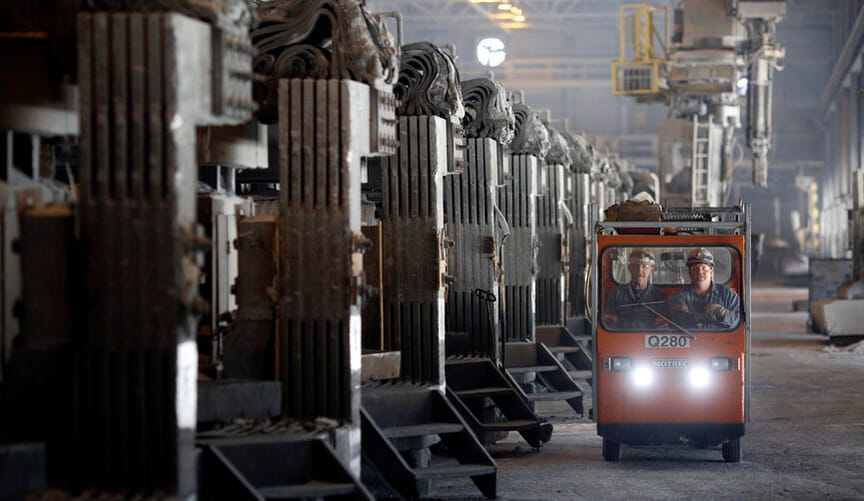 Two workers in safety gear ride in a small motorized vehicle inside a large factory