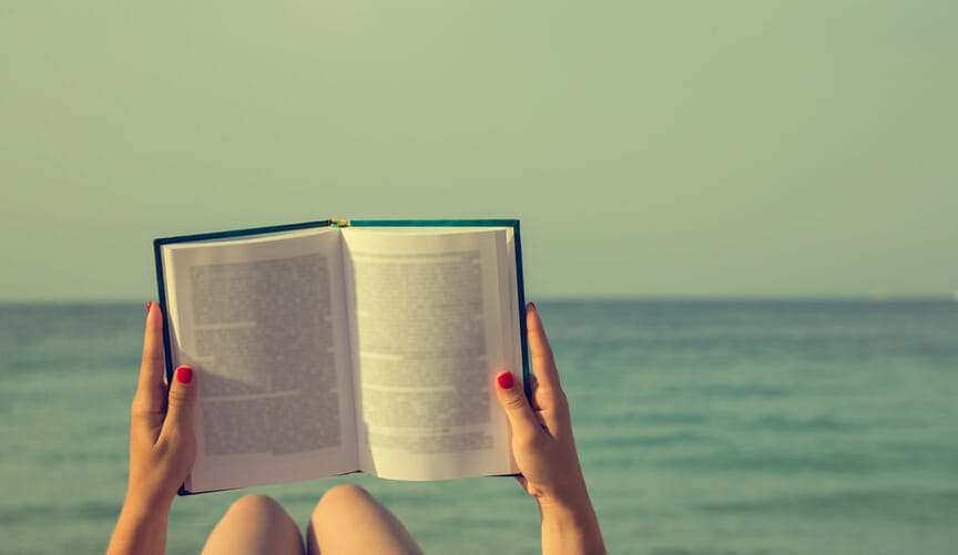 A person's legs and arms are shown reading a book next to a body of water