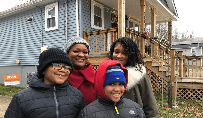 A Black woman and three Black children in winter coats smile in front of a small blue bungalow with holiday decorations