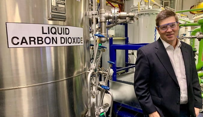 A white man in a dark sports coat and safety glasses stands in front of a large metal tank that says liquid carbon dioxide