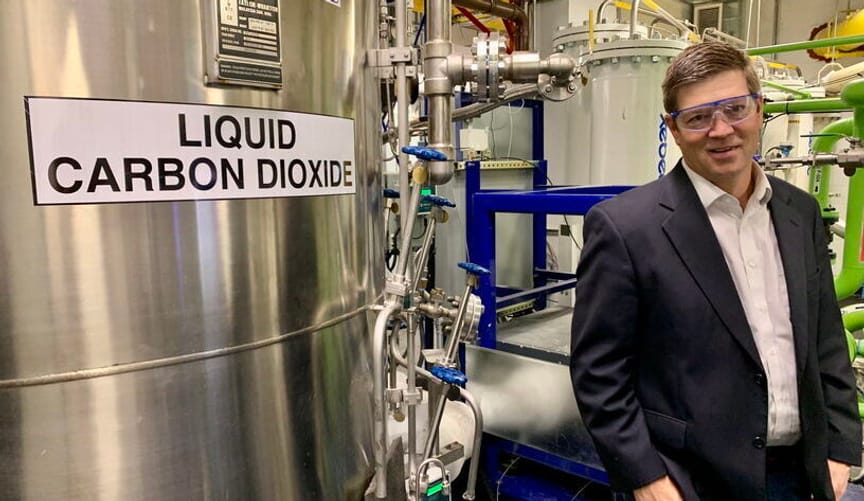 A white man in a dark sports coat and safety glasses stands in front of a large metal tank that says liquid carbon dioxide