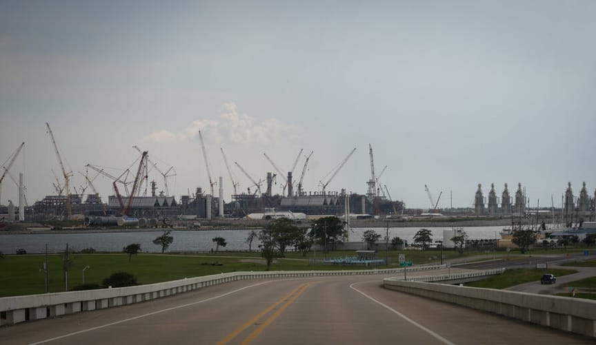 A road is in the foreground. In the distance is a huge industrial facility being built, with cranes jutting into the sky.