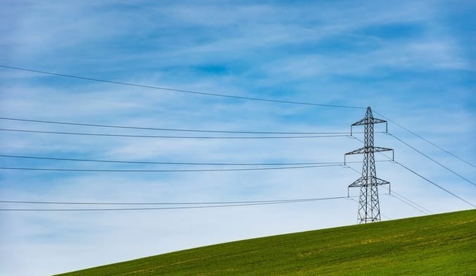 A lone black transmission tower on a grass covered hill against a blue sky with wispy white clouds