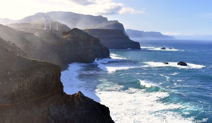 Ocean waves crash against a craggy coastline; wind  turbines can be seen on the cliff's edge in the distance