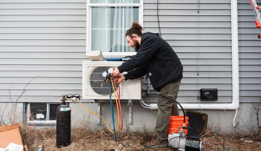 A man with a beard and ponytail hooks hoses to an appliance installed in an exterior window of a home