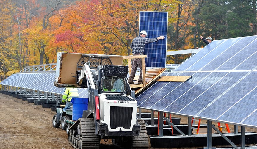 A worker installs solar panels on an array that is situated next to trees with bright autumnal foliage