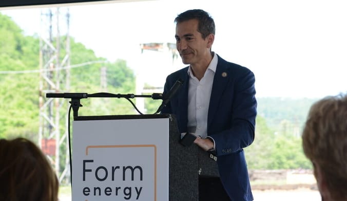 A man with a light skin tone and short dark hair wearing a blue blazer speaks at a lectern with a sign that says Form Energy