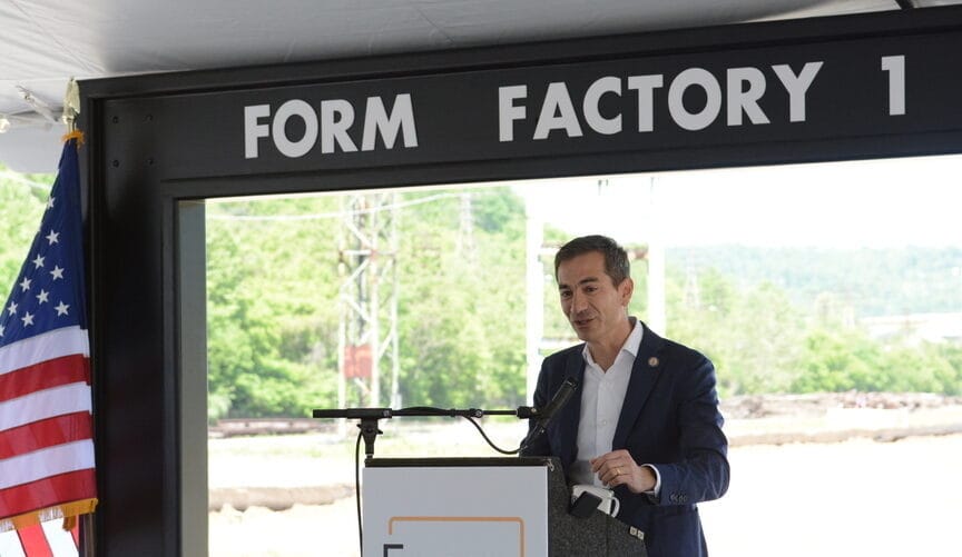 A man in a blue blazer and white shirt speaks at a lectern with a U.S. flag nearby under a sign that says form factory one