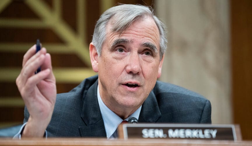 A man with a light skin tone and gray hair wearing a business suit speaks and gesticulates. A placard reads "Sen. Merkley."