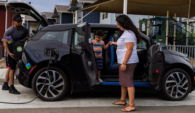 A woman with long dark hair helps a small boy get out of a black car, while a man wearing a baseball cap unloads the trunk