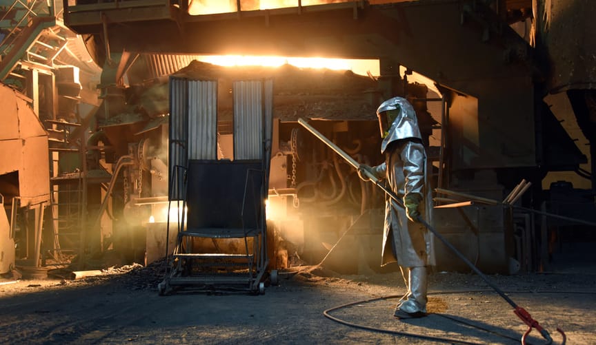 A worker in a full suit of protective gear oversees a blazing smelter inside a dark factory