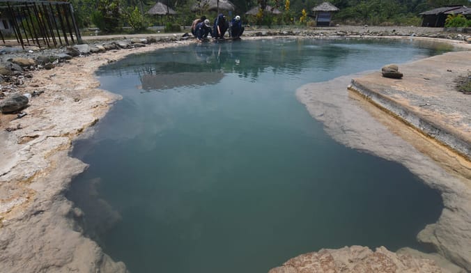 In the foreground is a small deep teal pond. Workers crouch on the far shore taking samples.