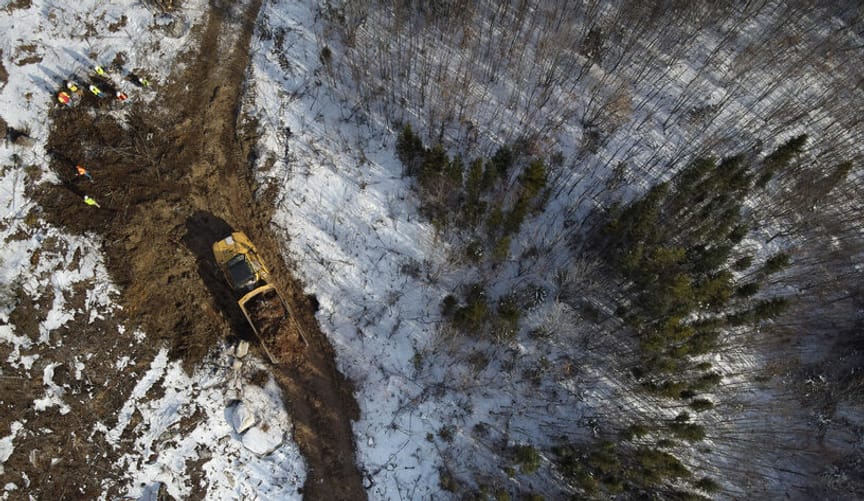 A tractor and several workers in safety gear are shown from above in a snowy, forested tract of land.