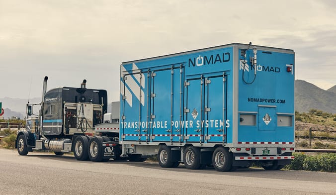 A semi truck pulling a blue trailer that says nomad transportable power systems through a desert landscape