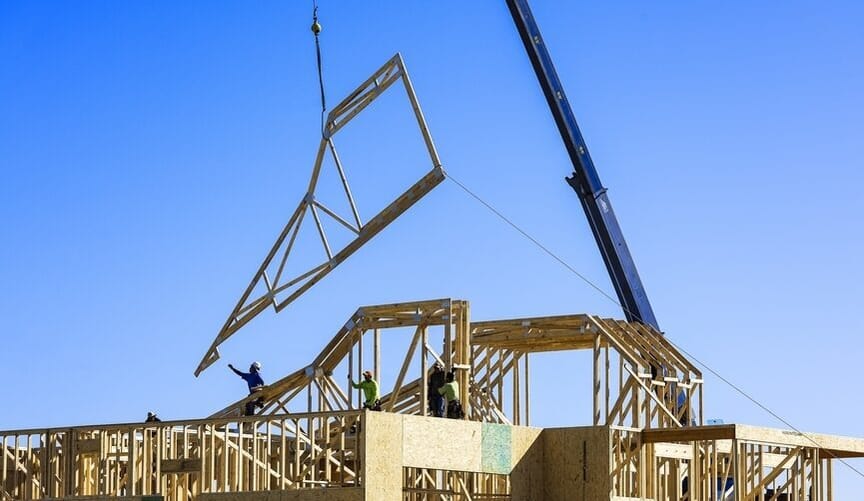Workers direct a large crane as it maneuvers a prefabricated panel of wood onto a home being constructed under a blue sky