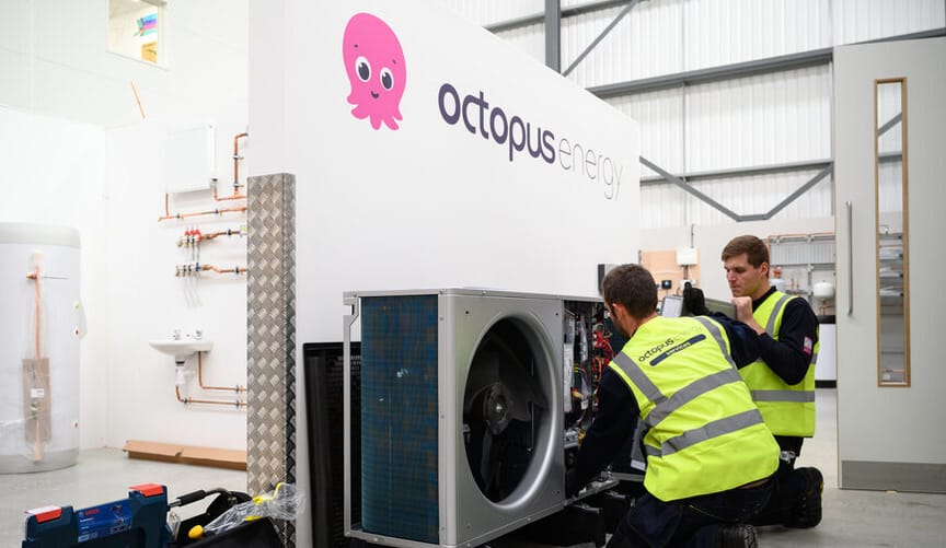 Two men in safety vests work on a heat pump in an indoor training center under a sign that says Octopus Energy