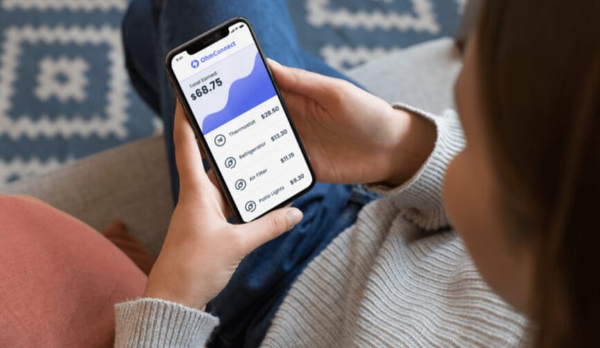 A woman sits on a couch looking at a smartphone with a message on energy savings from company OhmConnect